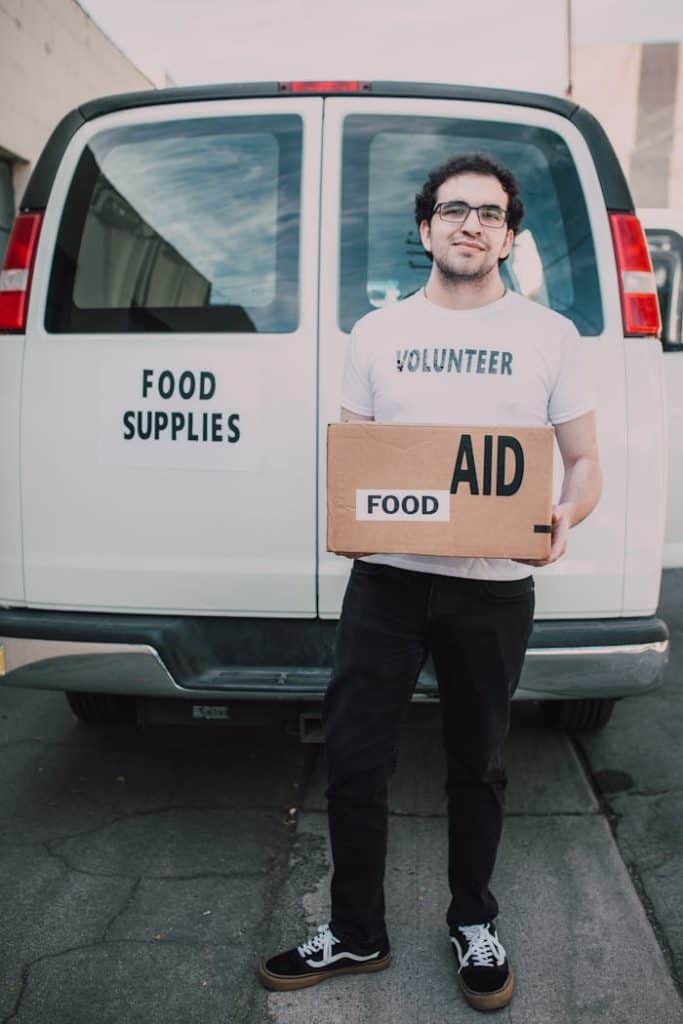 Man Wearing White Crew Neck Volunteer T-shirt Holding a Food Labelled Cardboard Box Behind White Van